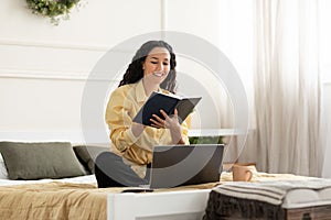 Smiling woman sitting on bed, using pc writing in notebook
