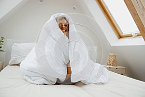 Smiling woman is sitting on a bed wrapped in a white blanket