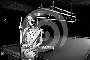 Smiling woman sitting on billiards table with cue.