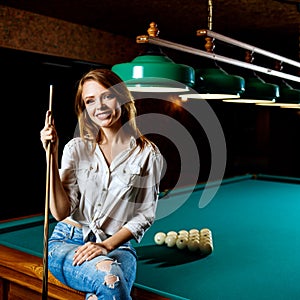 Smiling woman sitting on billiards table with cue.