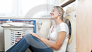 Beautiful smiling woman sitting on floor at laundry room and listening to music