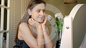 Smiling woman sitting at makeup table in room decorated with flowers. Skin care, female health and beauty