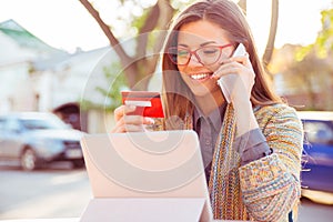 Smiling woman sitting outdoors talking on mobile phone making online payment on her tablet computer