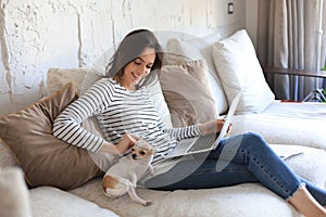 Smiling woman sitting on sofa with laptop computer, little dog playing near