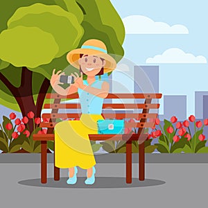 Smiling woman sitting on wooden bench and making selfie. Green tree, blooming flowers and city buildings on background