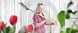 Smiling woman with spade in flower garden  on white wooden shed background with gardening tools and copy space