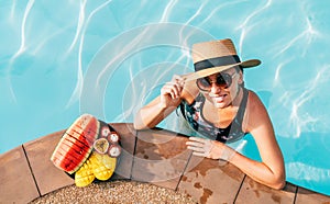 Smiling woman in straw hat in sunglasses swimming in pool and enjoying fresh tropical fruits