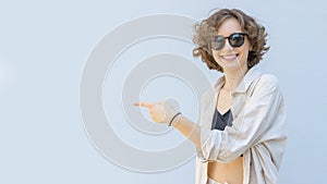 Smiling woman in sunglasses over isolated gray background pointing finger to the left side.