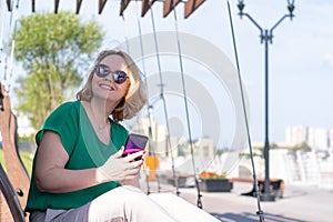 A smiling woman in sunglasses, talking on the phone with friends, making a date in the park in the summer.