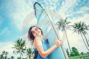 Smiling woman with surfboard posing on tropical beach.Surfer girl in big sunglasses with long board posing on the ocean beach.