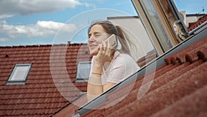 Smiling woman talking on her phone while gazing out of an open attic window, taking in the picturesque sight of red