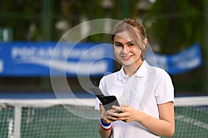 Smiling woman tennis player taking rest after training and using mobile phone. Sport, training and active life concept