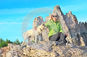 Smiling Woman Traveler and white dog sitting on stones relaxing with Rocky Mountain