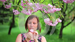 Smiling woman under blossomed sakura tree picks flower and decorates hair