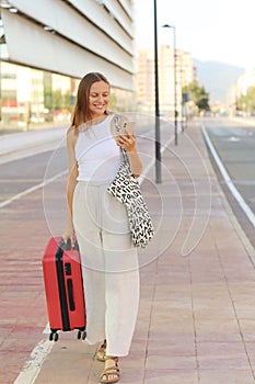 smiling woman uses cellphone while walking with red suitcase by the railway station or airport. Happy traveling girl