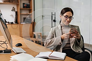 Smiling woman using cellphone while working with computer and planner