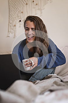 Smiling woman using laptop whilst in bed drinking mug of tea