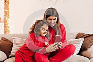 Smiling woman using smartphone with little daughter. Indoor shot of young mom and kid in red sitting on sofa