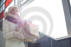 Smiling woman in warm clothing carrying stacked gifts by window