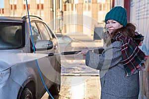 Smiling woman in warm wear washing her suv car at self car-wash station outdoors, using high pressure water jet