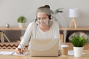 Smiling woman wearing headset writing notes studying online on laptop