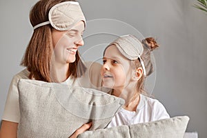 Smiling woman wearing sleeping mask sitting at home with her cute little daughter on bed talking looking at each other expressing