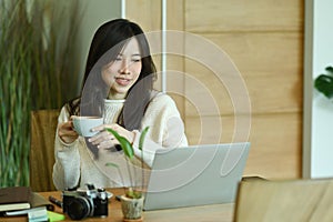 Smiling woman wearing warm sweater and drinking hot coffee while using laptop computer a home.