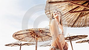 Smiling woman in a white bikini under straw beach umbrella, concept of a summer beach holiday, vacation travel and tourist resort