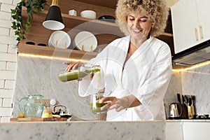 Smiling Woman in White Robe Pouring Green Juice in Kitchen