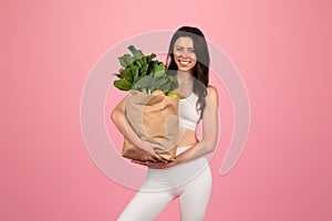 A smiling woman in a white sports outfit confidently holds a grocery bag full of green vegetables and fruits