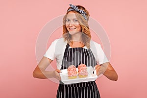 Smiling woman in white T-shirt and black apron showing fresh cakes, looking at camera with happy expression, delicious pastry,