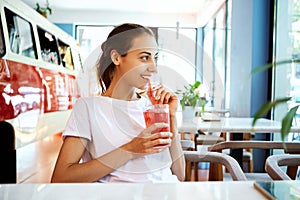 Smiling woman in white T-shirt drinking red berry lemonade or cocktail and looking out the window. Young pretty female