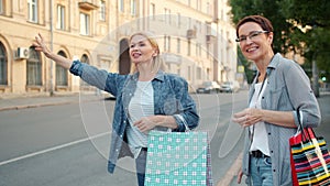 Smiling women with shopping bags hailing taxi waving hand outdoor in city street