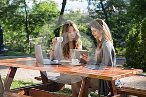 Smiling women sitting outdoors in park drinking coffee using laptop