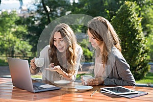 Smiling women sitting outdoors in park drinking coffee using laptop