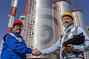 Smiling Worker and Businessperson Shaking Hands Against Oil Refinery Storage Tanks