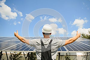 Smiling worker with solar station, raising his hands, showing thumbs up on a background of photovoltaic panels near the