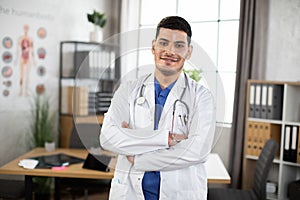 Smiling young 30-aged male Arabic Asian doctor posing with arms crossed in the office