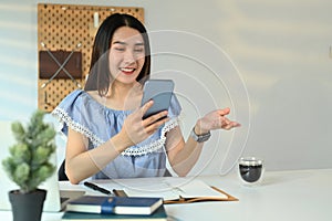 Smiling young adult asian woman chatting with client online on smart phone and using laptop at her workplace