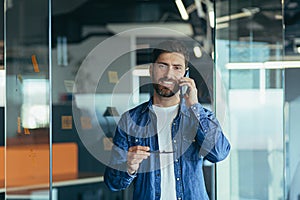 Smiling young adult bearded hipster professional business man making a business call while talking on the phone in the office