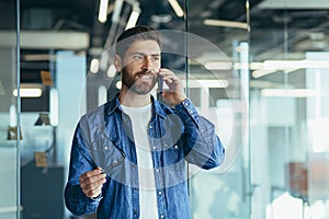 Smiling young adult bearded hipster professional business man making a business call while talking on the phone in the office