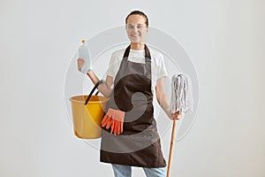 Smiling young adult woman housewife wearing white t shirt and brown apron holding equipment for cleaning flat, mop, ppduster and