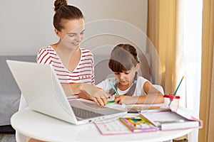 Smiling young adult woman wearing striped casual clothing helping her little daughter to do homework, distance education during