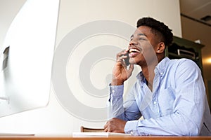 Smiling young african american businessman talking on phone while looking at computer