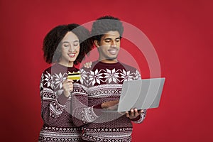 Smiling young african american couple in identical Christmas sweaters, guy typing on laptop