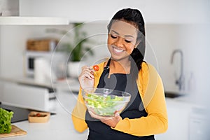 Smiling young african american female chef in apron eating organic vegetable salad, enjoy fresh dish