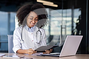 Smiling young African American female doctor working in clinic office with laptop and documents, sitting at table
