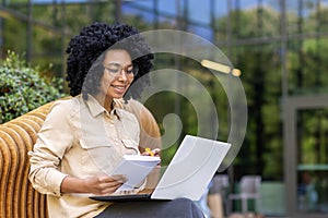 Smiling young African American female student sitting on a bench on a university campus studying online using a laptop