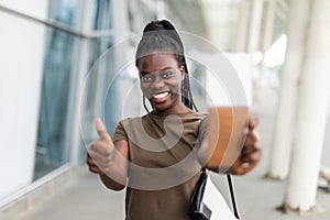 Smiling young african girl hold passport boarding pass tickets showing thumb up in airport terminal