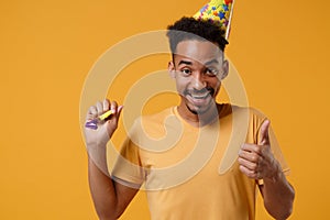 Smiling young african american guy in casual t-shirt, birthday hat posing isolated on yellow orange wall background